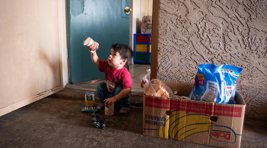 Young boy sitting outside his home with a food box holding up a food item