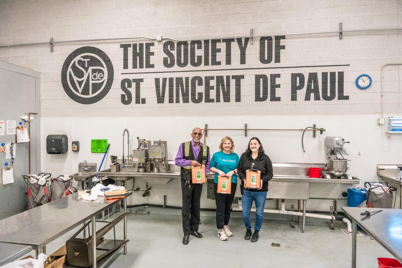 Two member of the security team stand with a volunteer, all holding donated AEDs, in front of the SVdP seal and logo.